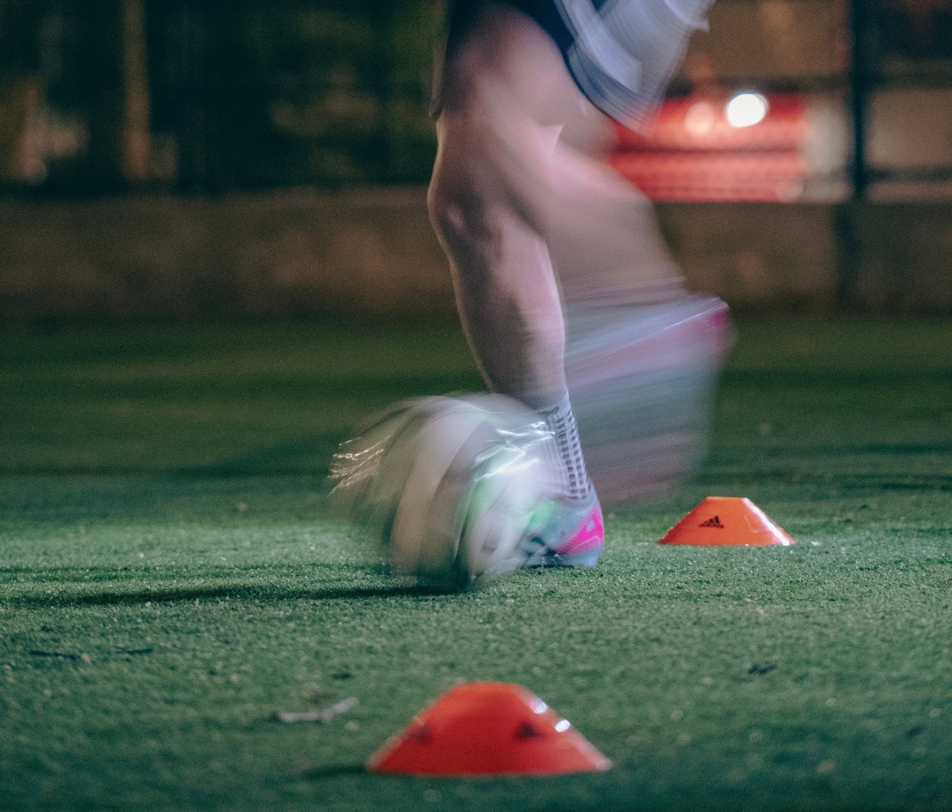 Soccer players foot kicking a soccer ball on artificial turf field at night with training cones nearby