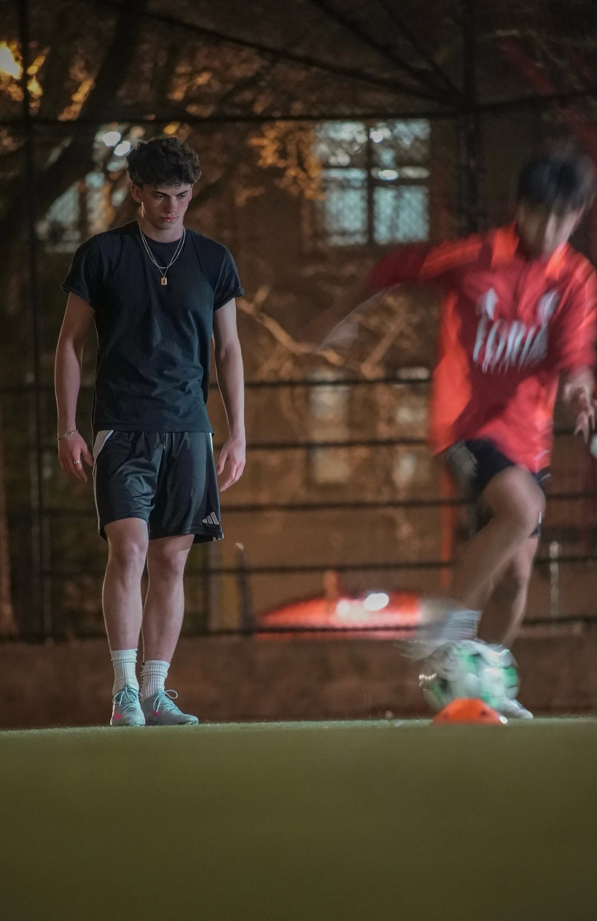 Young soccer player in dark athletic wear standing on field watching teammate in red jersey during training session in evening light